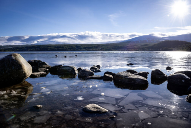 Photograph of loch with a foreground of large stones in frozen water with sheets of ice, with snow covered mountains in the distance